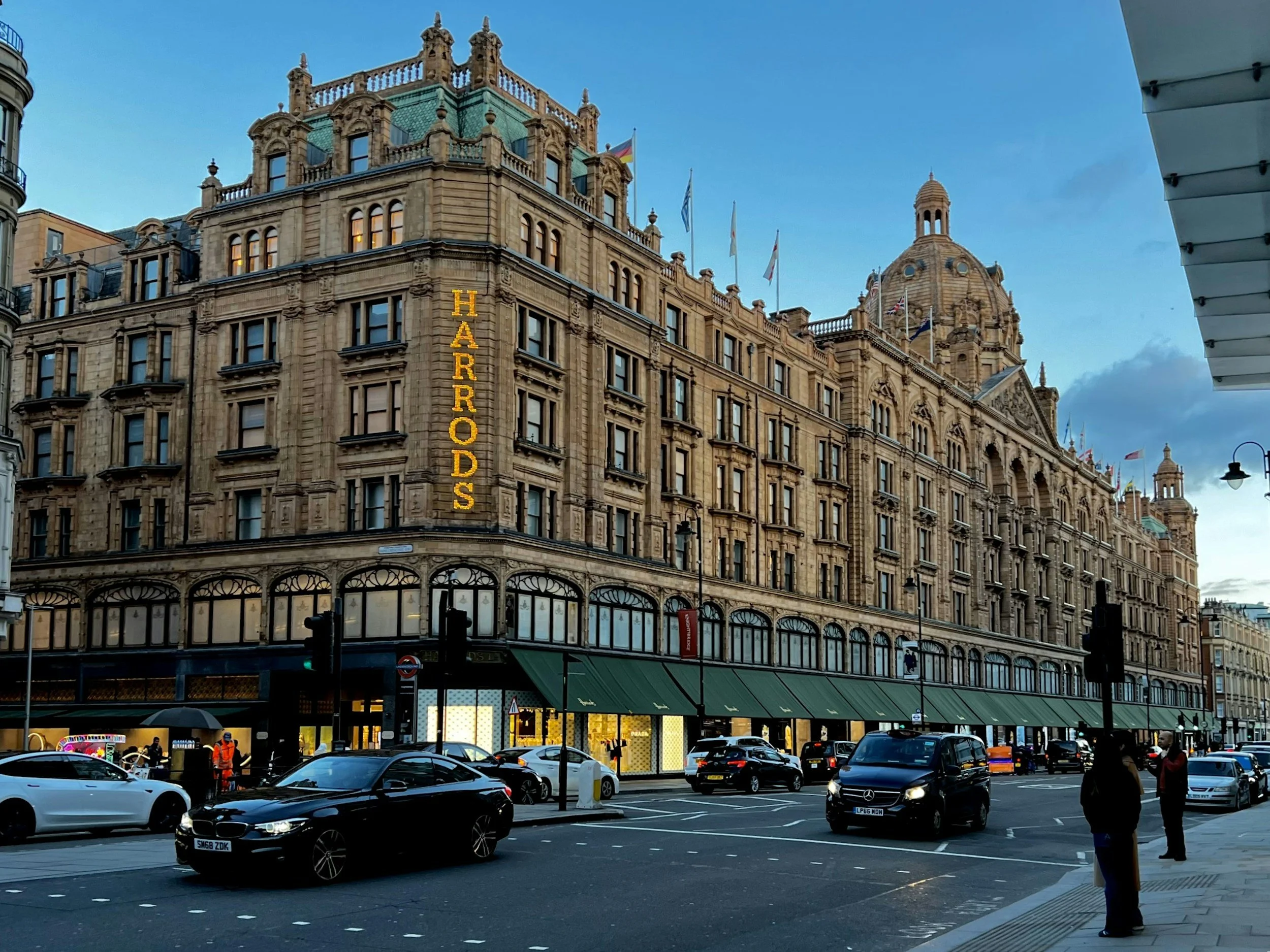 A large historic hotel with a green roof and ornate architecture called Harrods, located in London. The street in front has cars and people, with a clear evening sky in the background.