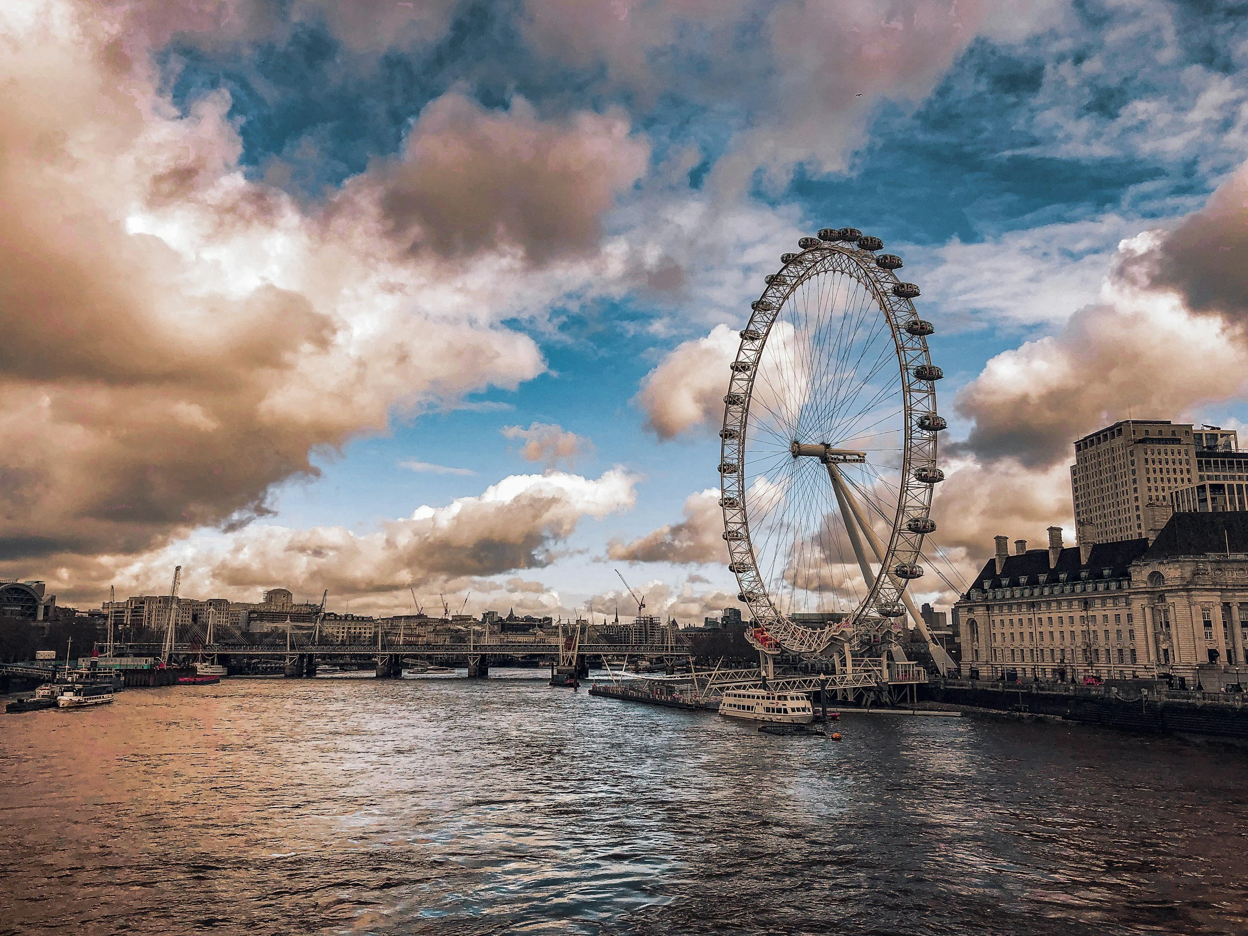 View of the London Eye Ferris wheel on the River Thames under a partly cloudy sky during sunset, with buildings along the riverbank.