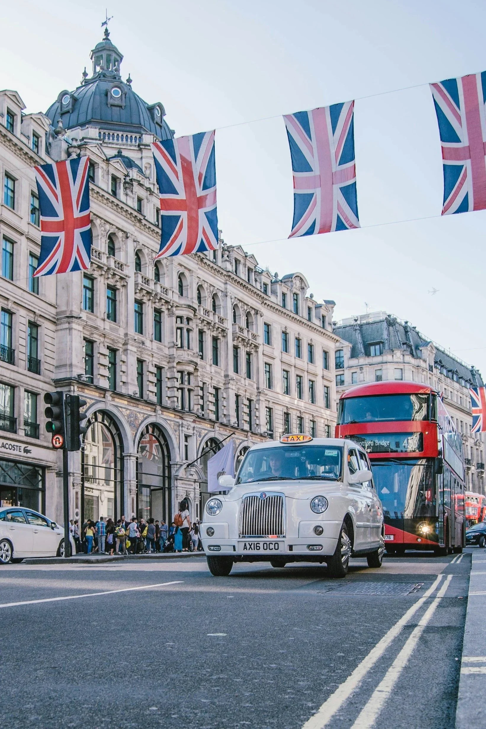 Street scene in London with Union Jack flags hanging and classic taxis, double-decker buses, and historic buildings.
