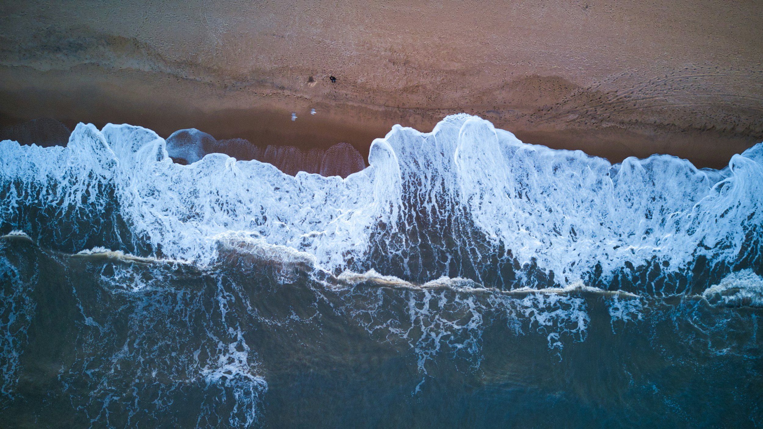 Aerial view of ocean waves crashing onto sandy beach with footprints and two people visible from above.