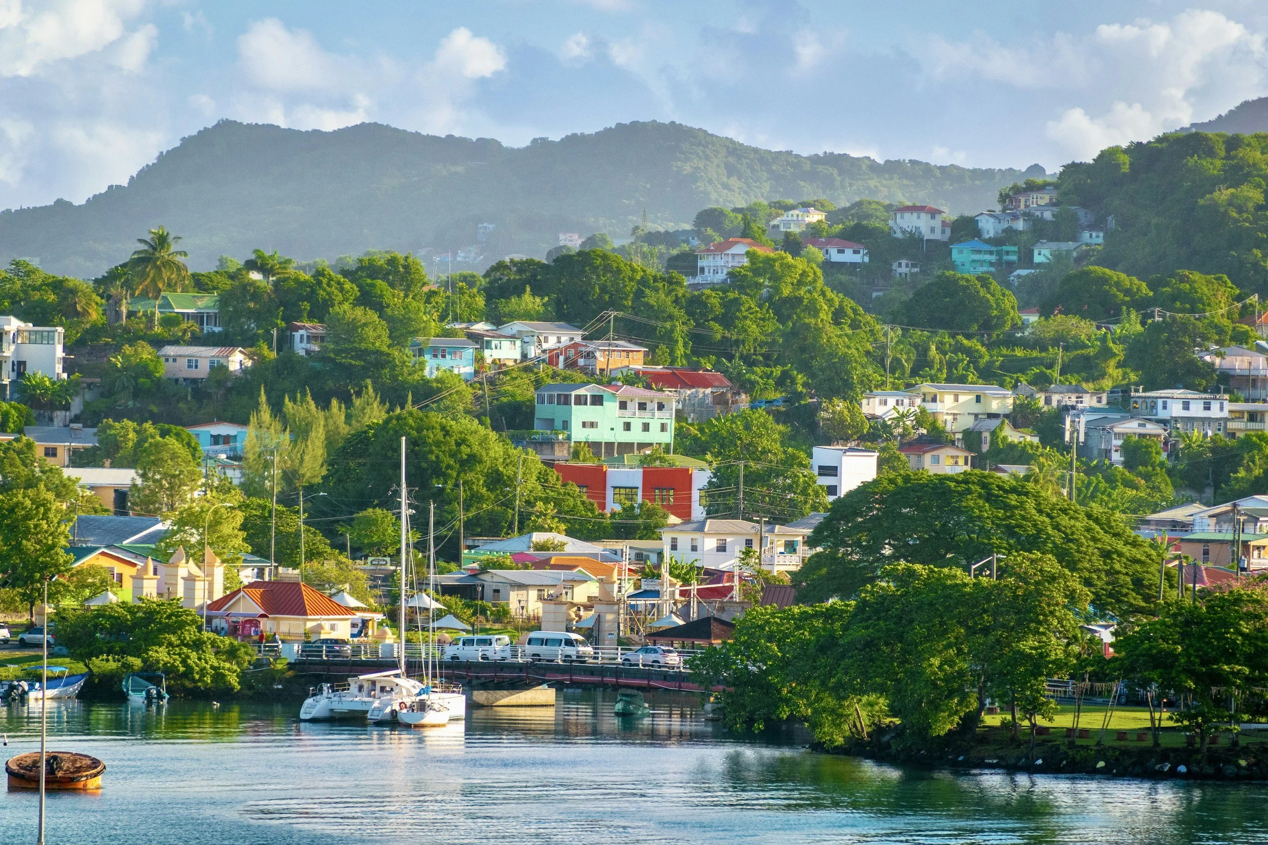 A colorful hillside town with houses and buildings, a body of water in the foreground, boats docked near the shore, and lush green trees, with mountains in the background under a partly cloudy sky.