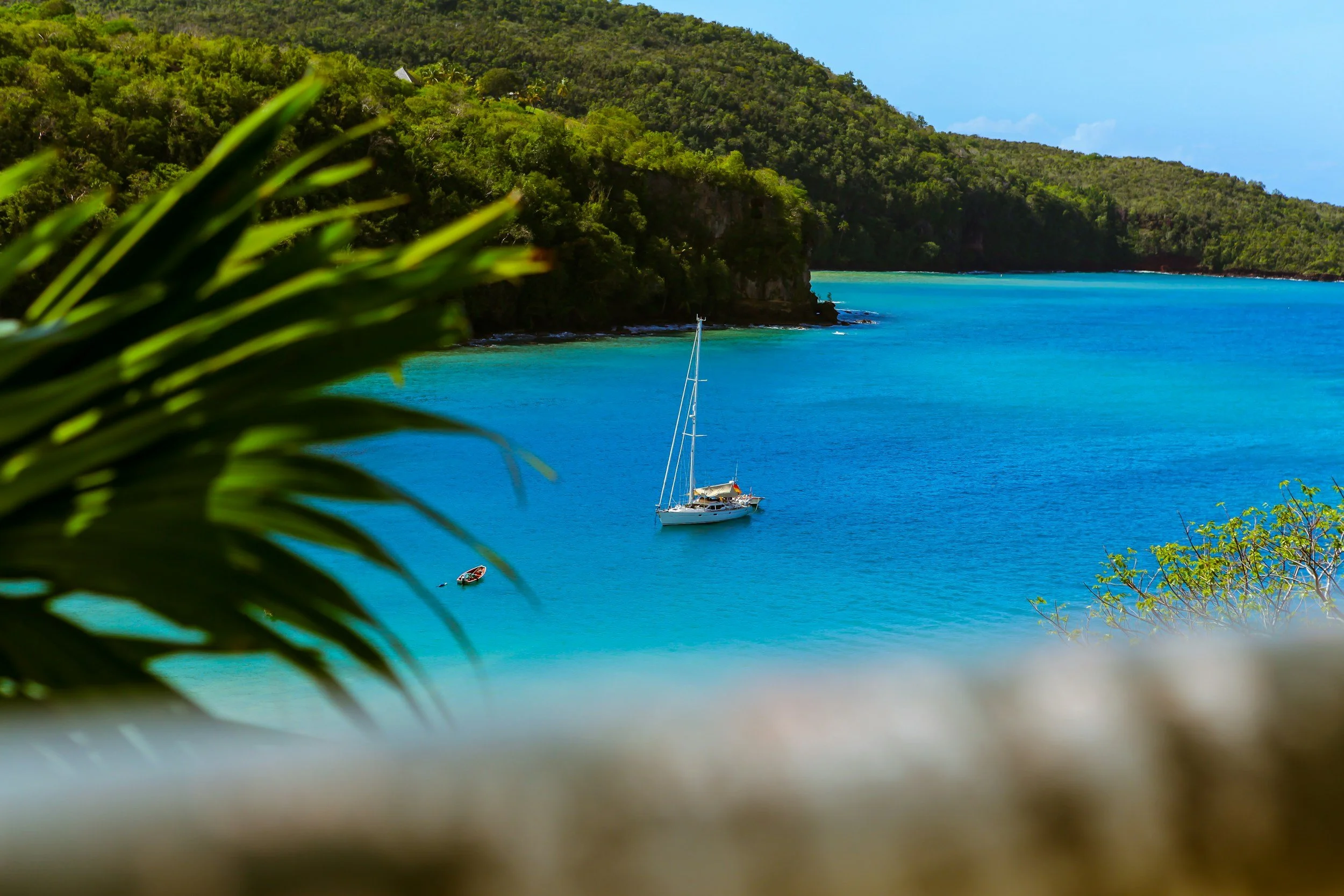 Tropical bay with turquoise water, green-covered hills, a sailboat, and a small boat, seen through tropical plants in the foreground.