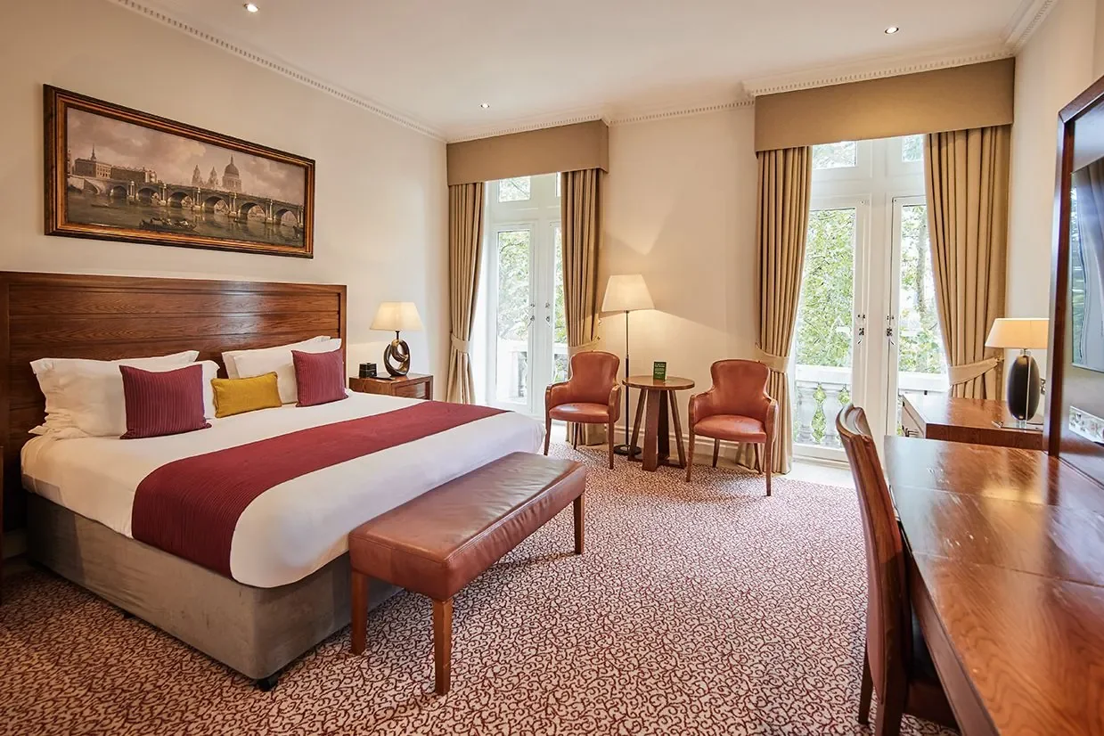 A hotel room with a large bed, two chairs and a small table, large windows with beige curtains, a dresser with a mirror, and artwork depicting a bridge and cityscape on the wall.