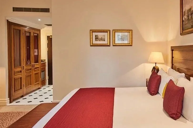 A cozy hotel room with a large bed featuring a red bed runner and matching pillows. There are framed pictures on the wall above the bed and a bedside lamp turned on, casting warm light. In the background, a wooden wardrobe and part of a small kitchen