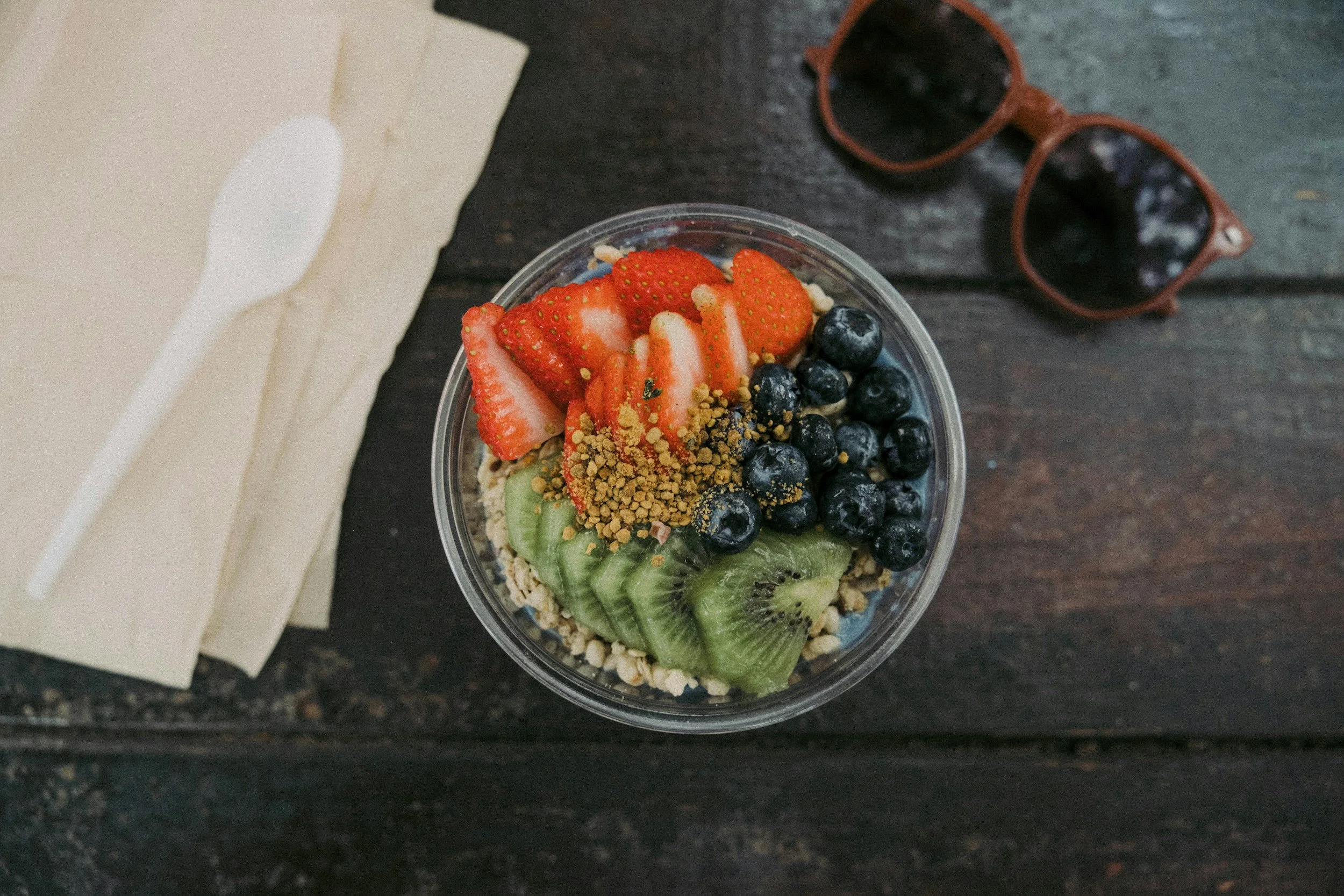 A cup of fruit and granola topped with sliced strawberries, blueberries, kiwi, and crushed nuts, placed on a wooden table with sunglasses and napkins nearby.