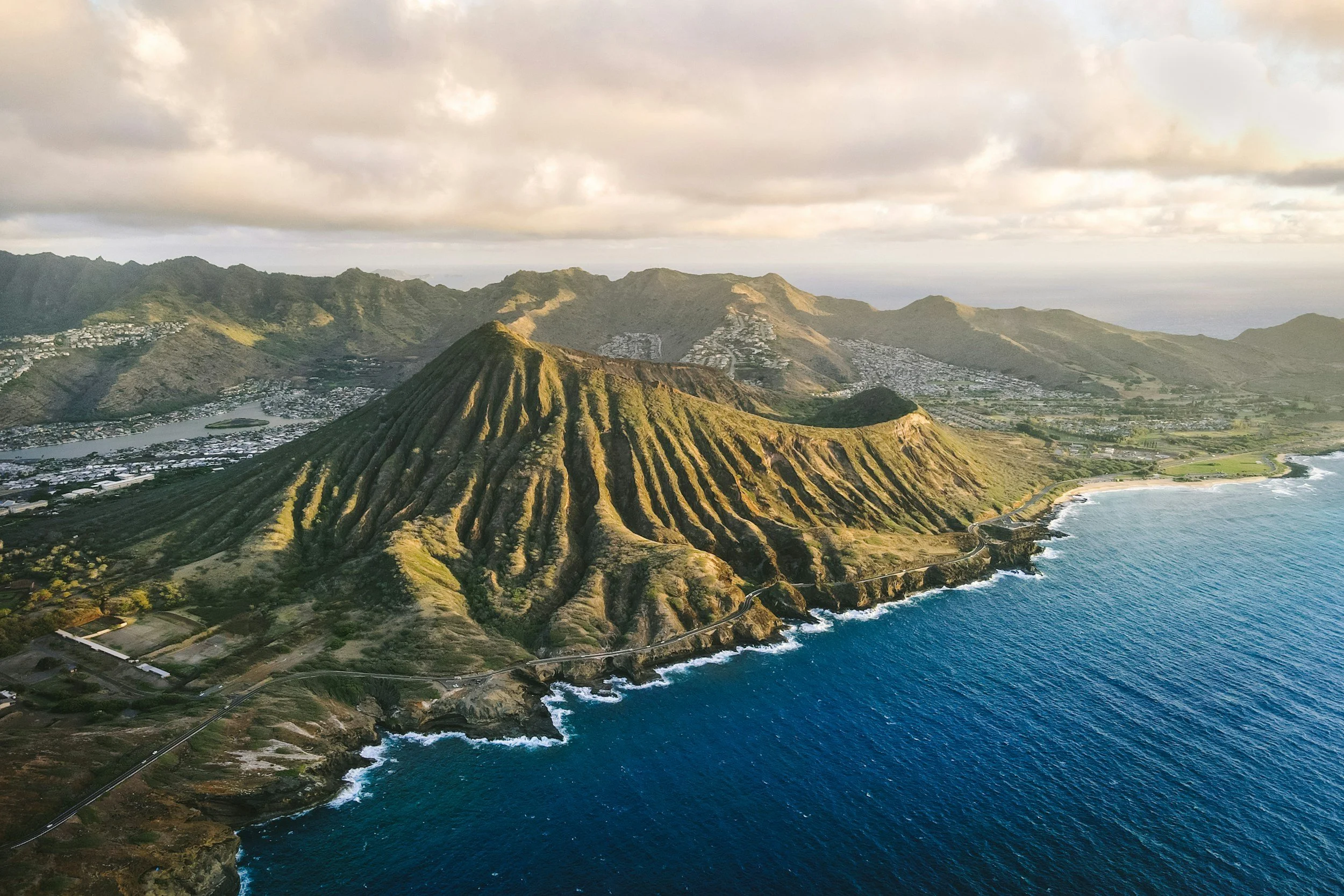 Aerial view of a rugged green volcano near the coastline, with a road running along its base, overlooking the ocean, under a cloudy sky.