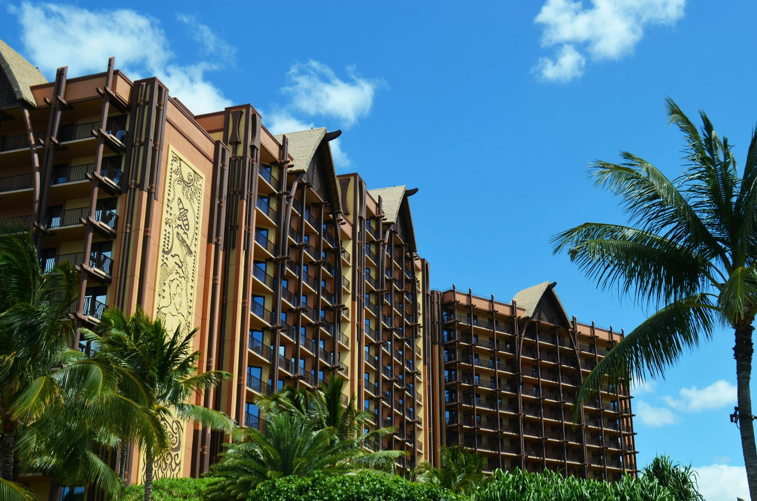High-rise resort building with balconies, surrounded by palm trees, against a bright blue sky with clouds.