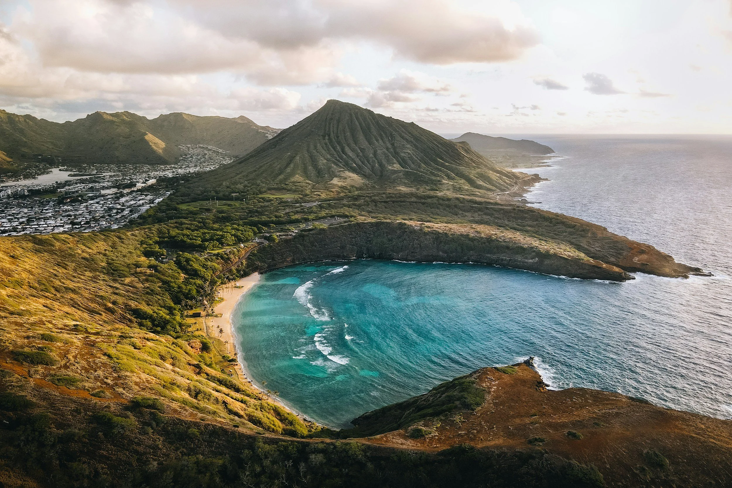 A coastal scene with a sandy beach, a crescent-shaped bay with turquoise waters, lush green hills, and a tall, conical volcanic mountain near the shoreline. Overcast sky with some clouds.