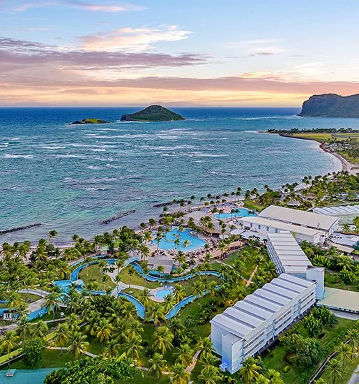 Aerial view of a coastal resort with multiple swimming pools, lush palm trees, and beachfront with ocean and small islands in the background at sunset.