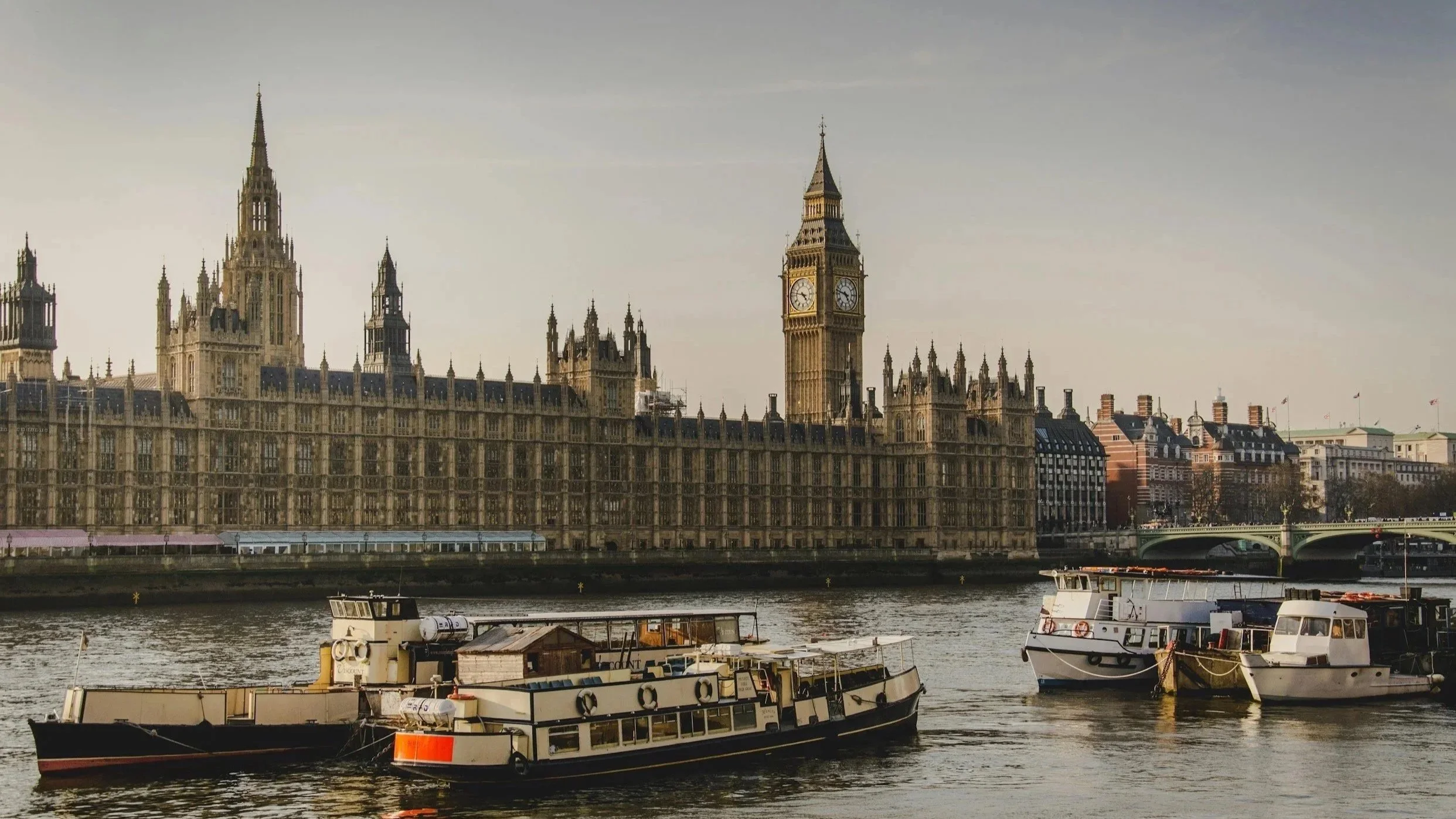 View of the Houses of Parliament and Big Ben in London, England, with boats on the river in the foreground during daytime.