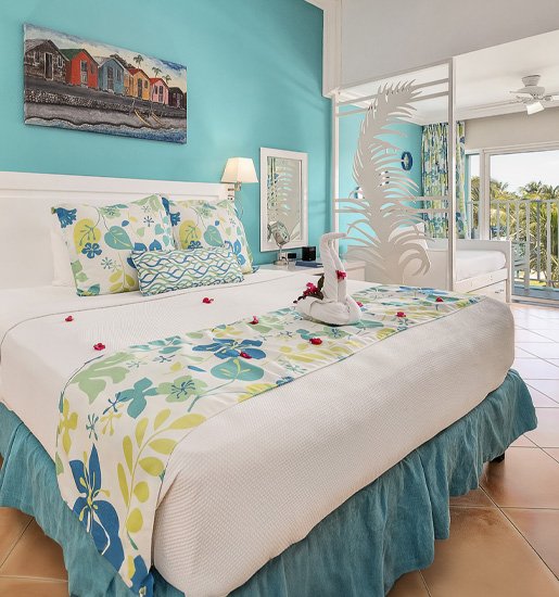 A bedroom featuring a white bed with a floral bed runner and pillows, decorated with a towel swan and rose petals, a blue wall with a colorful shoreline painting, a white sliding door, and a balcony with water views.