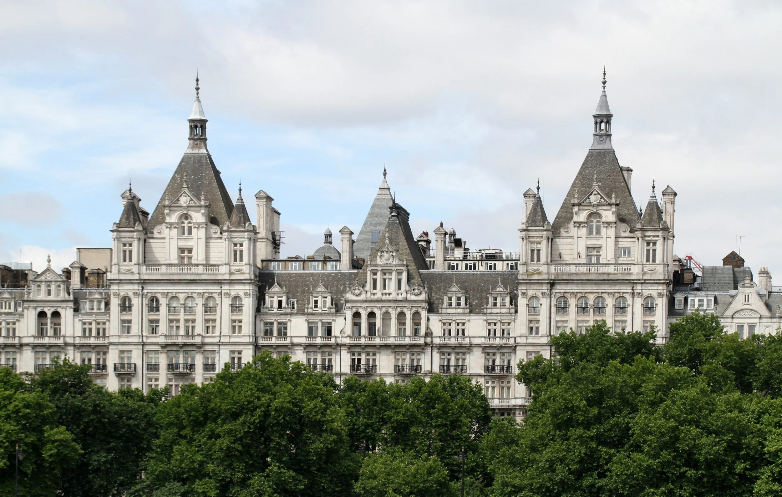 Historical castle-like building with gothic towers and ornate architecture, surrounded by green trees under a cloudy sky.
