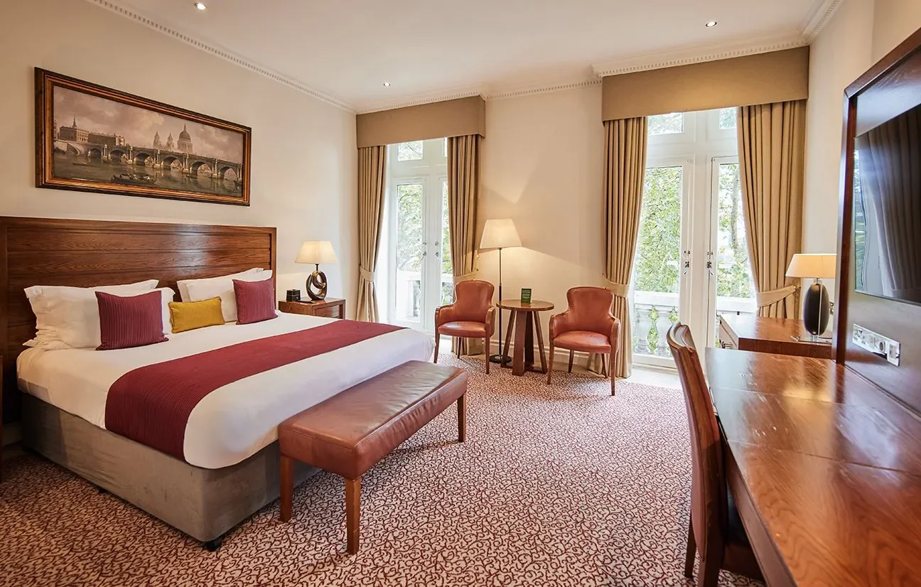 A hotel room with a large bed, two orange chairs, a small round table, and large windows with beige curtains, a wooden desk, and a framed painting of a bridge and buildings.