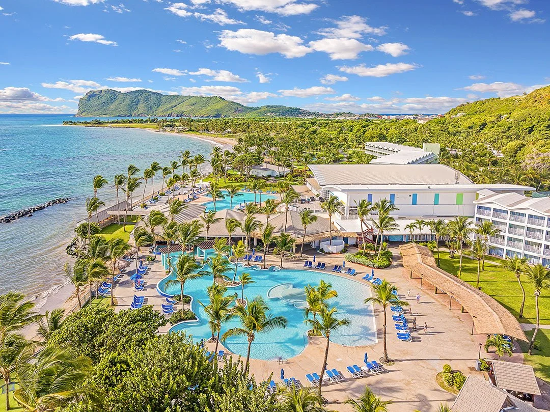 Aerial view of a tropical resort with multiple swimming pools, palm trees, and beachfront access, overlooking the ocean with a mountain in the distance.