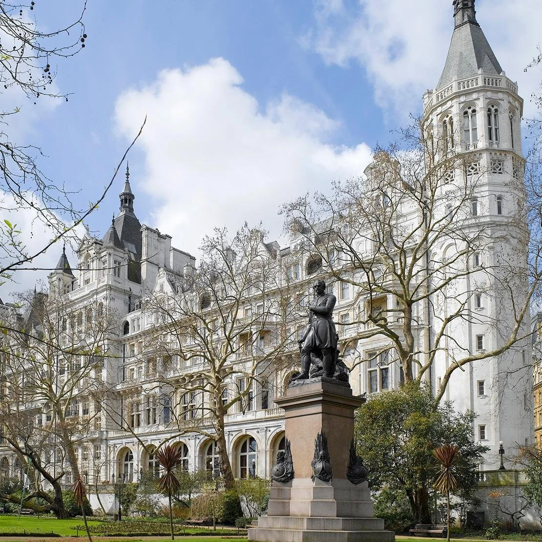 A historic white castle-like building with turrets and towers, surrounded by leafless trees, with a historic statue in the foreground in a park setting.