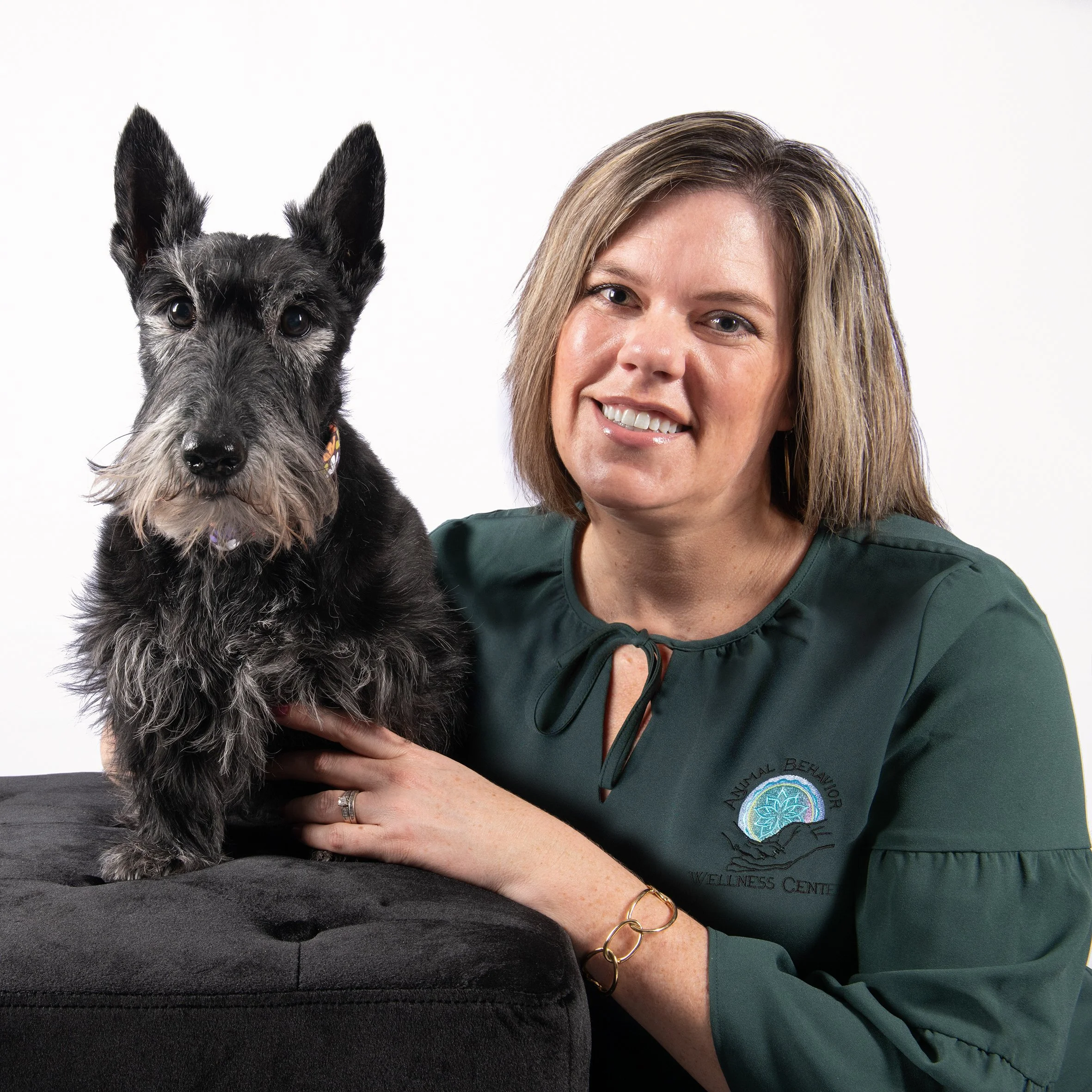 A woman with shoulder-length light brown hair smiling and wearing a dark green shirt with an embroidered logo, sitting next to a black and gray dog with wire hair and pointed ears, against a plain white background.
