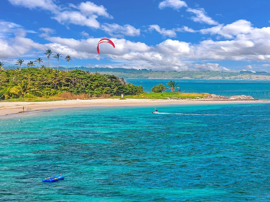 Tropical beach with turquoise water, a person jet skiing, a person walking on the sand, a red kite in the sky, and lush green trees with palm trees along the shoreline under a partly cloudy sky.