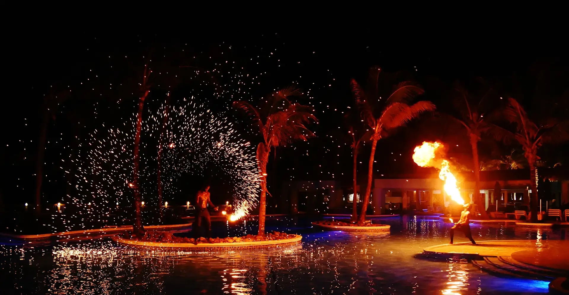 Night scene at a pool with two fire performers, one creating sparks and the other blowing fire, surrounded by palm trees and reflections in the water.