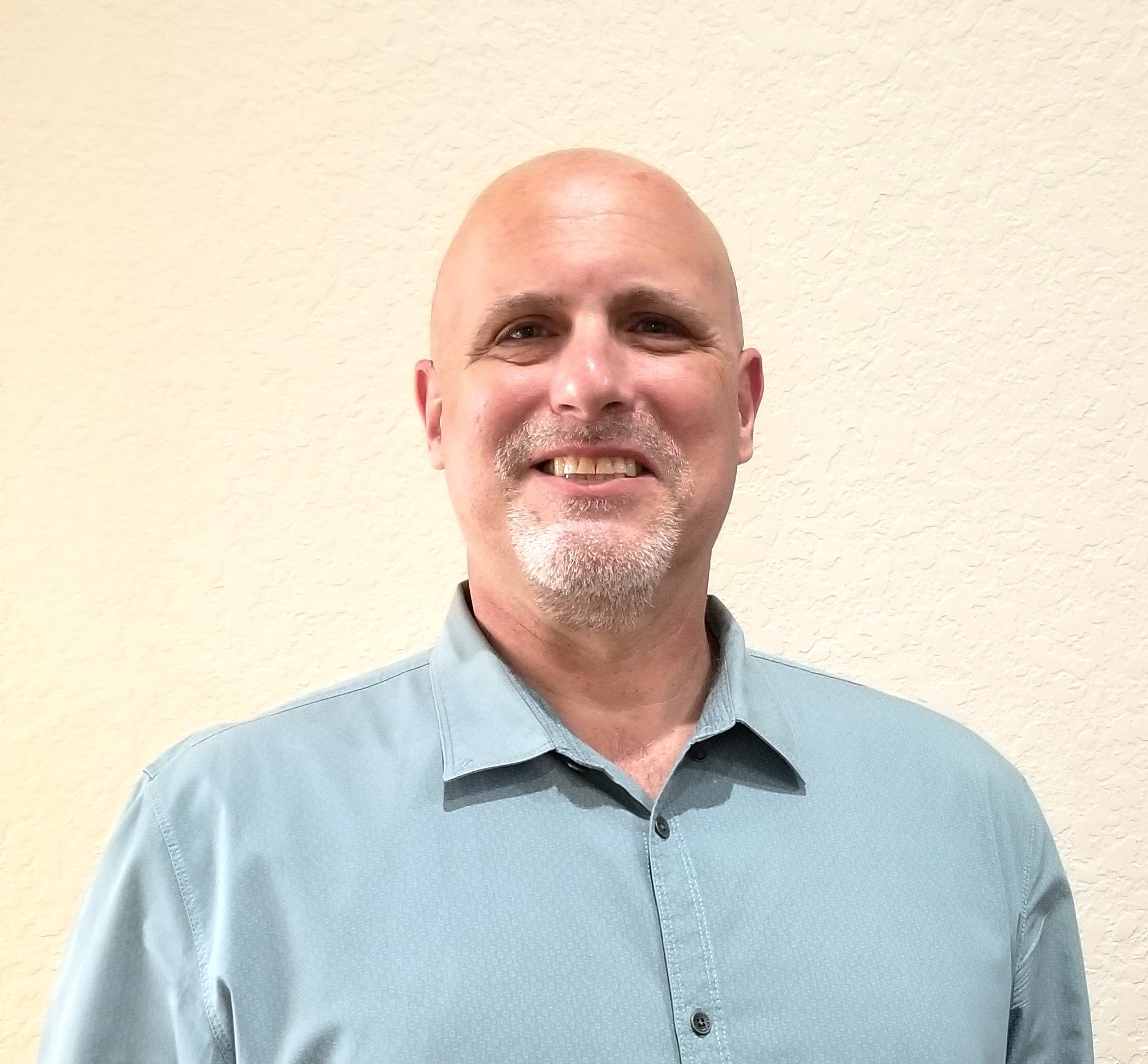 A middle-aged man with a goatee and bald head smiling, wearing a light blue collared shirt, standing against a beige textured wall.