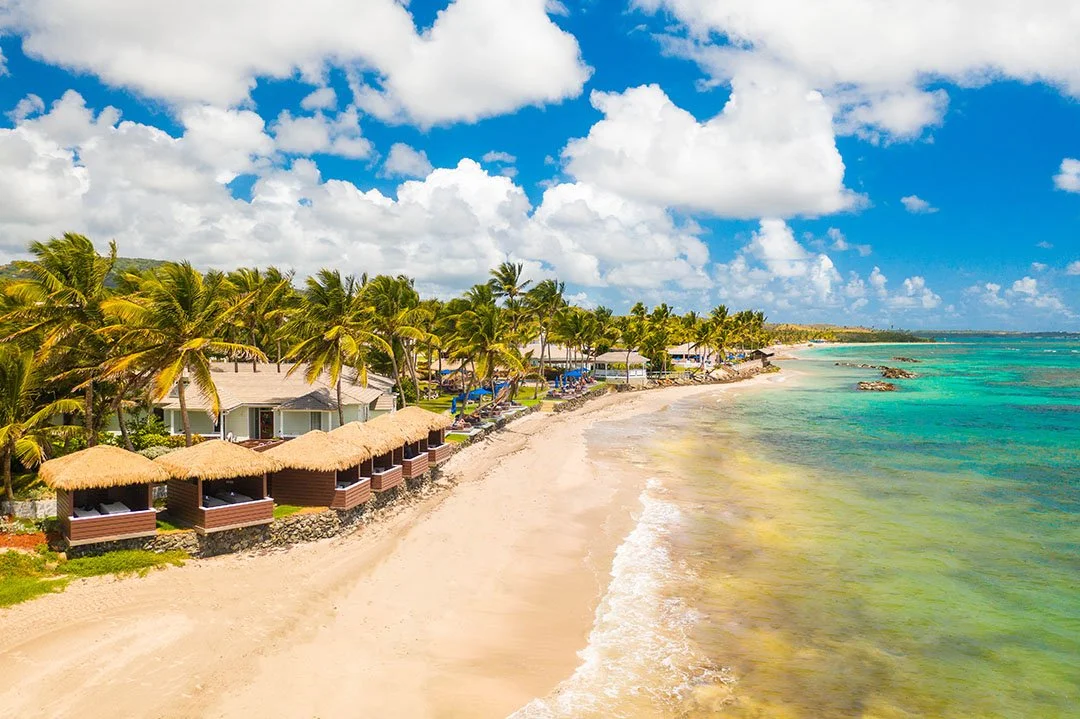 Tropical beach with white sand, turquoise water, and palm trees lining the shore. There are beachfront cabanas with thatched roofs and chairs, under a partly cloudy sky.