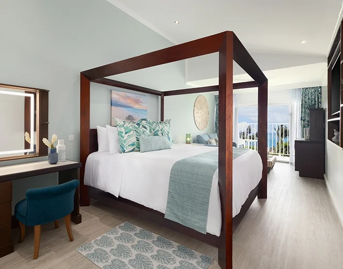 A bedroom with a wooden four-poster bed, white bedding, and teal decorative pillows, situated near a balcony with ocean views.