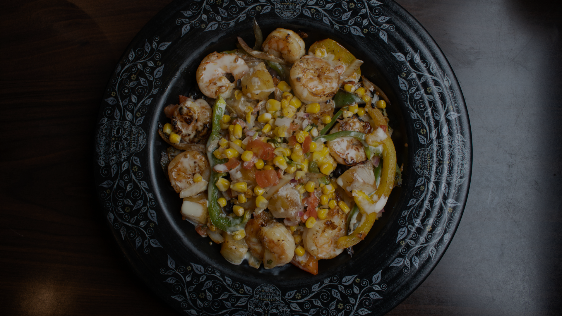 Plate of sautéed shrimp with yellow, green, and orange bell peppers, corn, and diced vegetables on a decorative black plate.