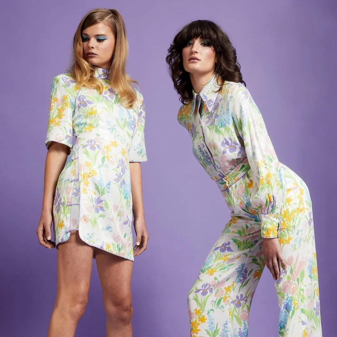 Two women wearing matching floral dresses, standing against a purple background.