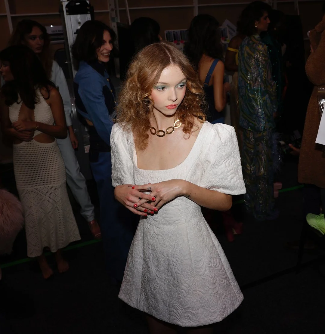 A woman with curly red hair wearing a white textured dress and a chunky gold necklace, standing among other women in a crowded room.