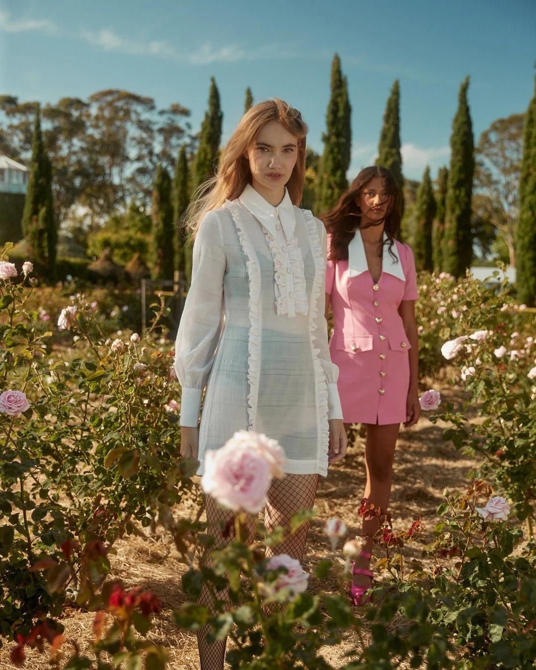 Two women standing among pink roses in a garden on a sunny day, one wearing a white dress with ruffles and fishnet stockings, the other in a pink dress with gold buttons.