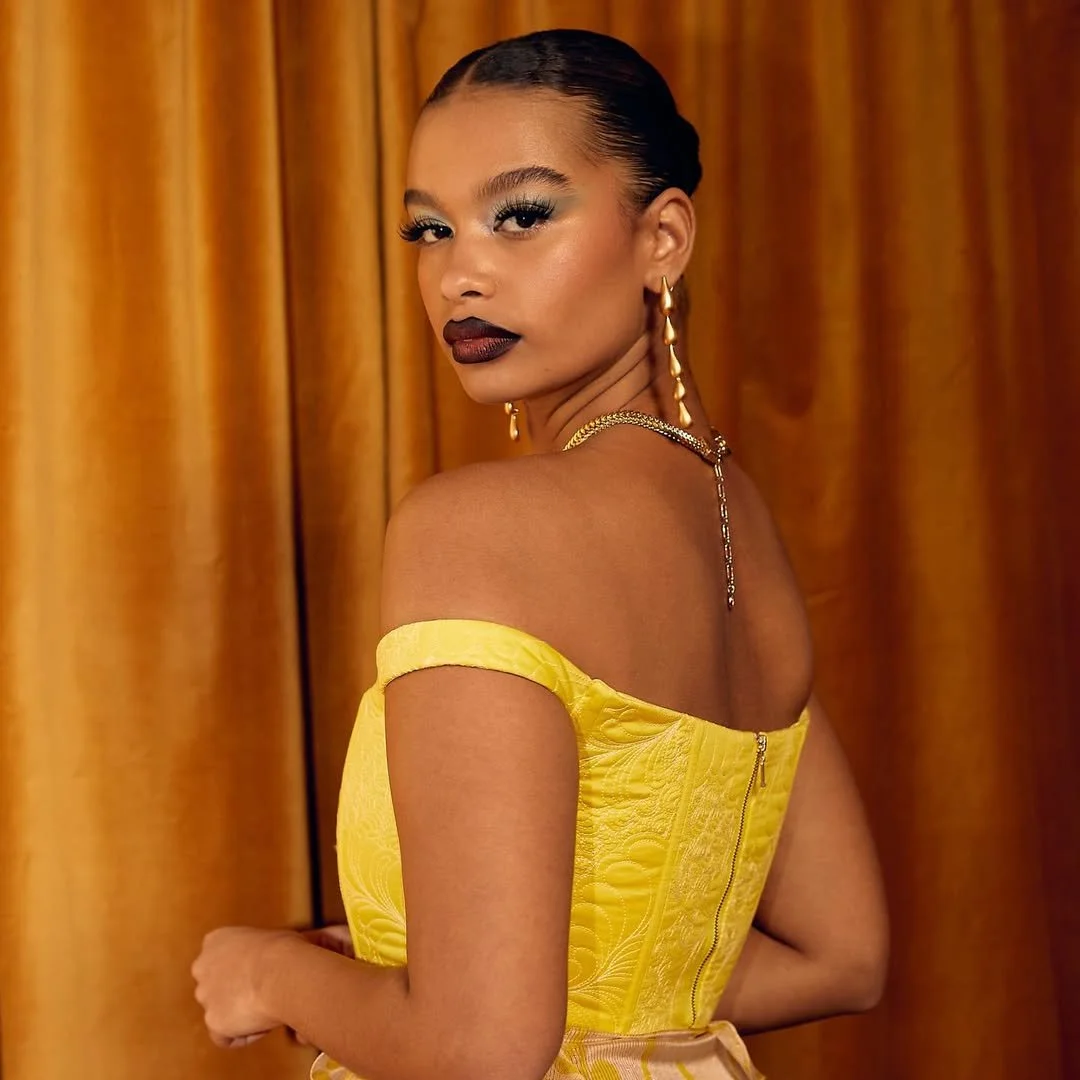 Young woman in yellow off-shoulder dress with dark lipstick and gold jewelry standing in front of gold curtains