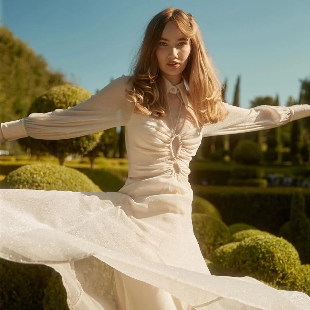 A young woman with long wavy hair wearing a cream-colored, long-sleeve blouse and a flowing skirt, outdoors in a sunny, well-maintained garden with pruned bushes and trees.