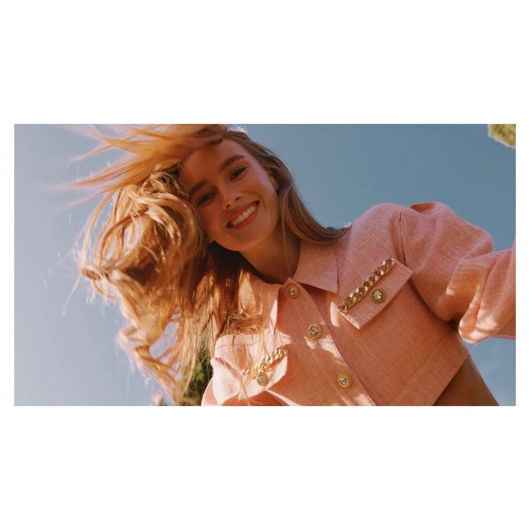 A smiling woman with long blonde hair wearing a pink coat with pearl and gold accents, looking down at the camera against a blue sky.