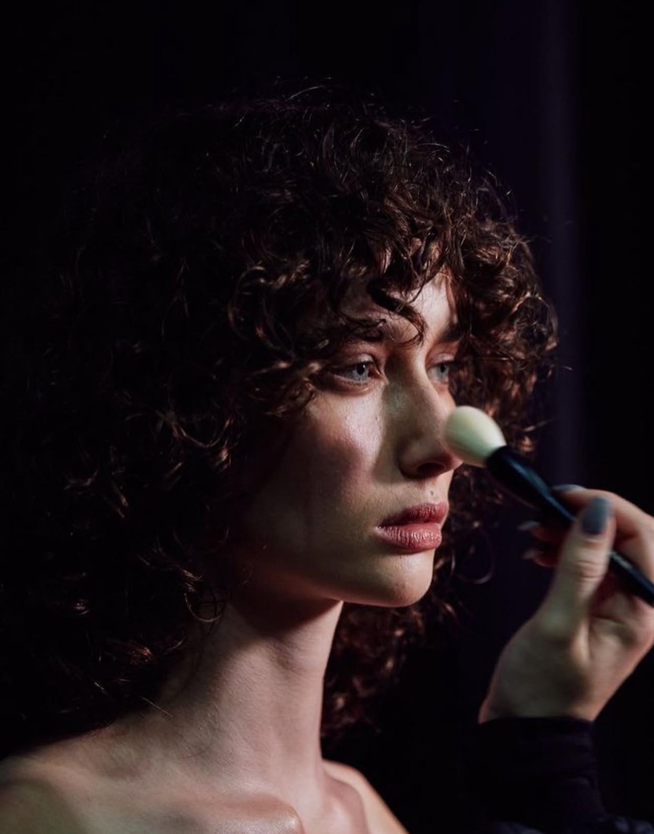 Close-up of a woman with curly hair applying makeup with a brush on her nose against a dark background.