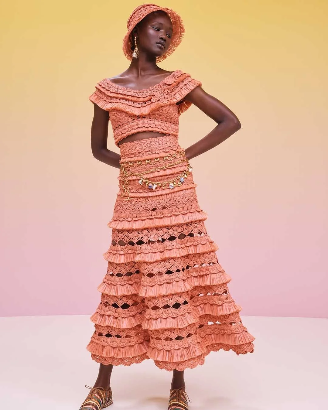 A woman dressed in a layered, coral-colored, crochet outfit with a matching hat, standing against a gradient background.