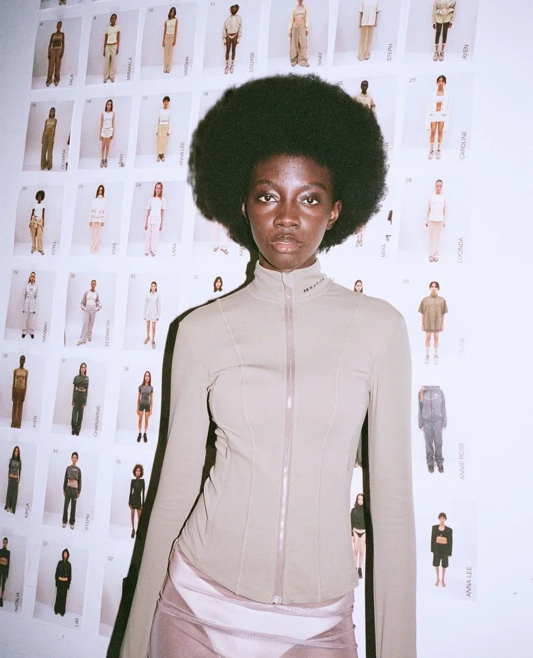 A woman with a large natural afro hairstyle standing in front of a wall covered with models' photographs and names. She is wearing a beige zip-up jacket.