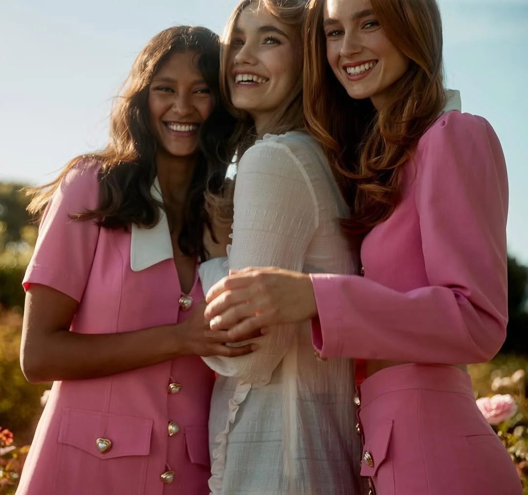Three women smiling and standing outdoors in bright sunlight, with two wearing pink outfits and one in a white dress.