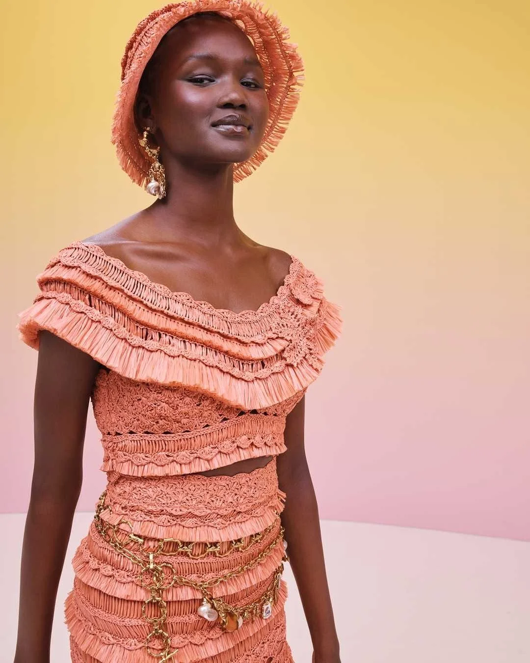 A woman wearing a peach-colored, ruffled, textured dress with decorative chains and pearls. She has matching earrings and a brimmed hat, standing against a gradient background of yellow, pink, and cream.