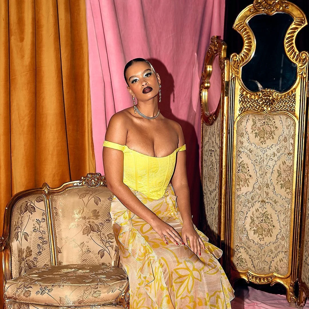 A woman sitting on an antique sofa in a room with colorful curtains, wearing a yellow off-shoulder top and a floral skirt, with jewelry and makeup.