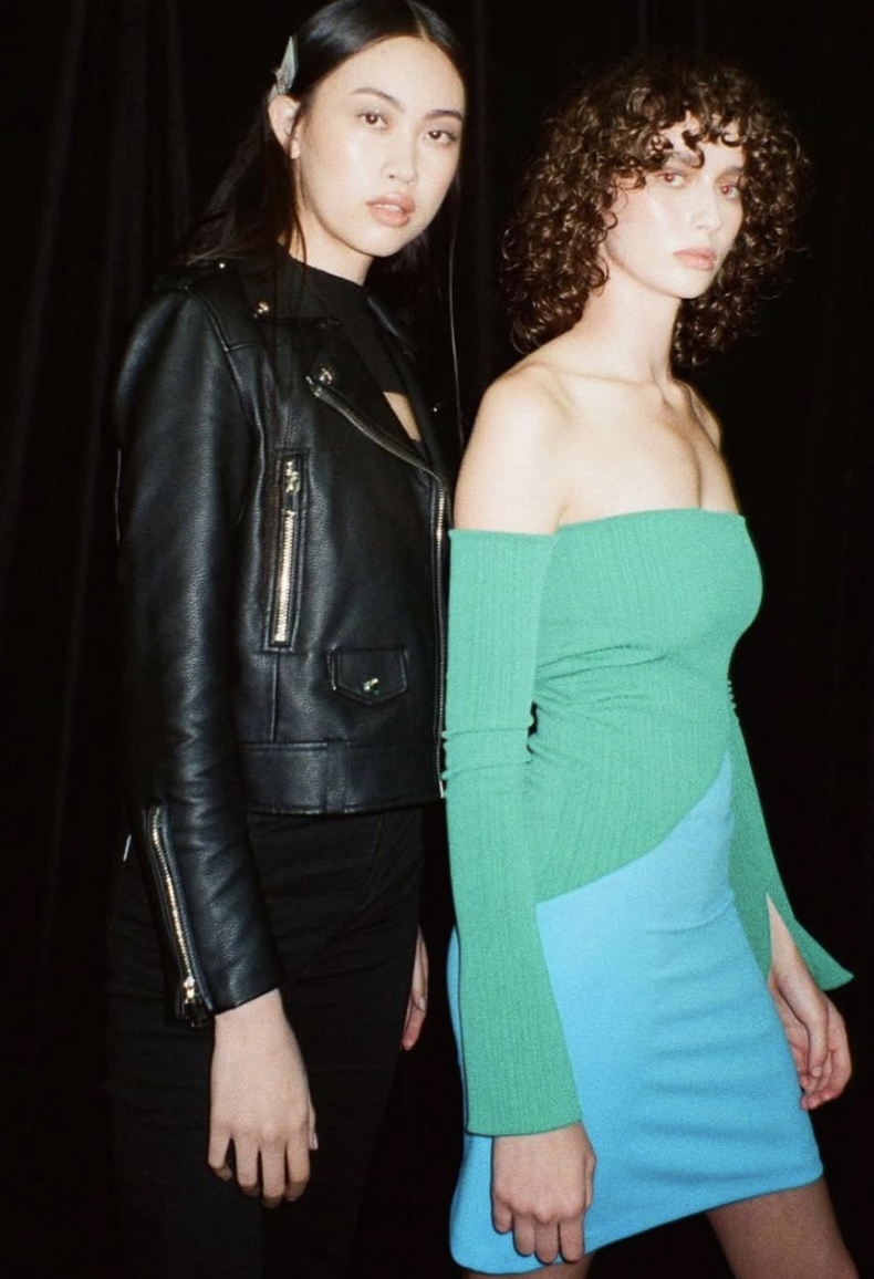 Two women standing together against a black background. The woman on the left has long dark hair, wearing a black leather jacket and black pants. The woman on the right has short curly hair, wearing an off-the-shoulder green top and a blue skirt.