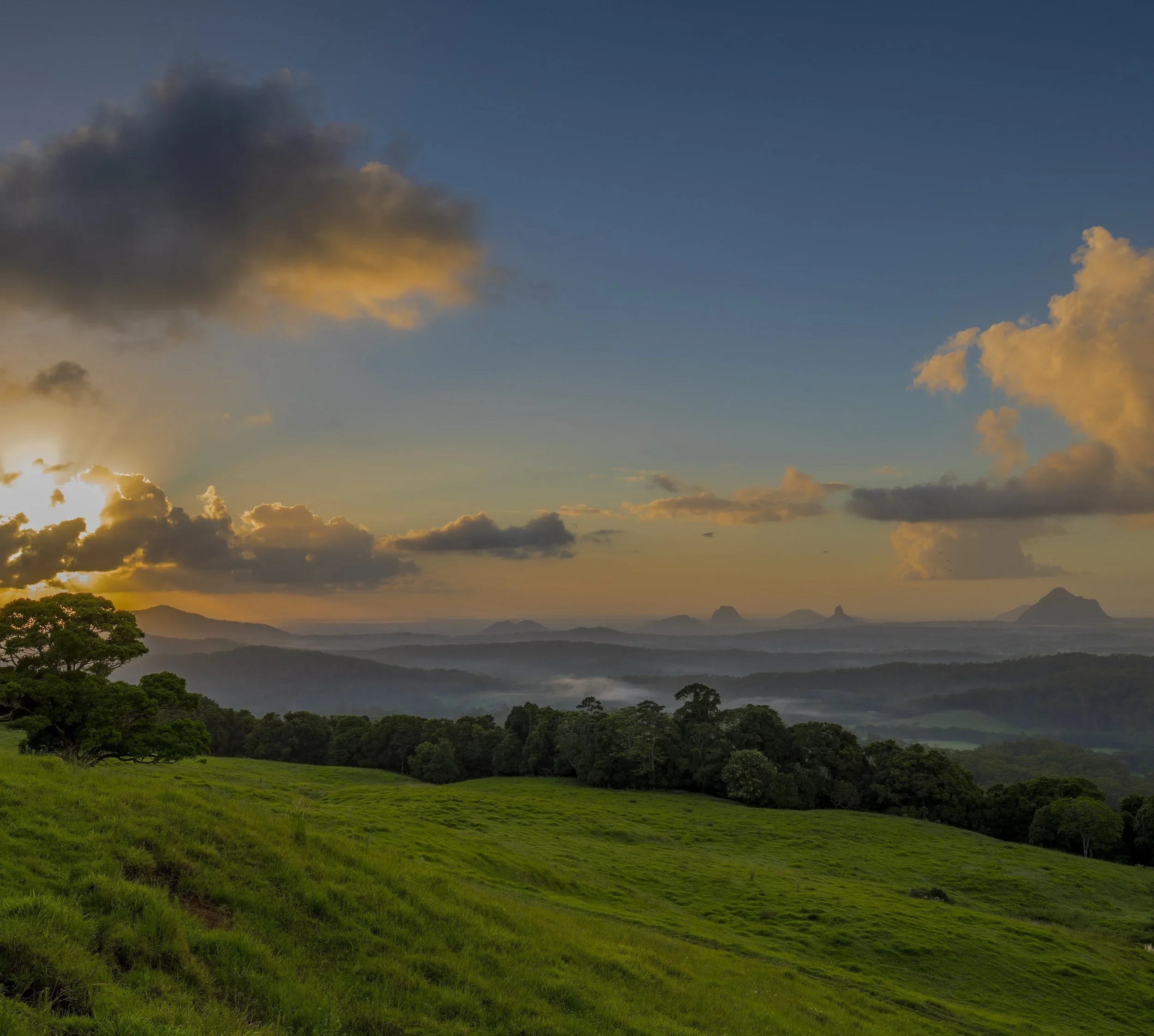 Sunset over a green hilly landscape with trees and distant mountains, cloudy sky with golden sunlight.