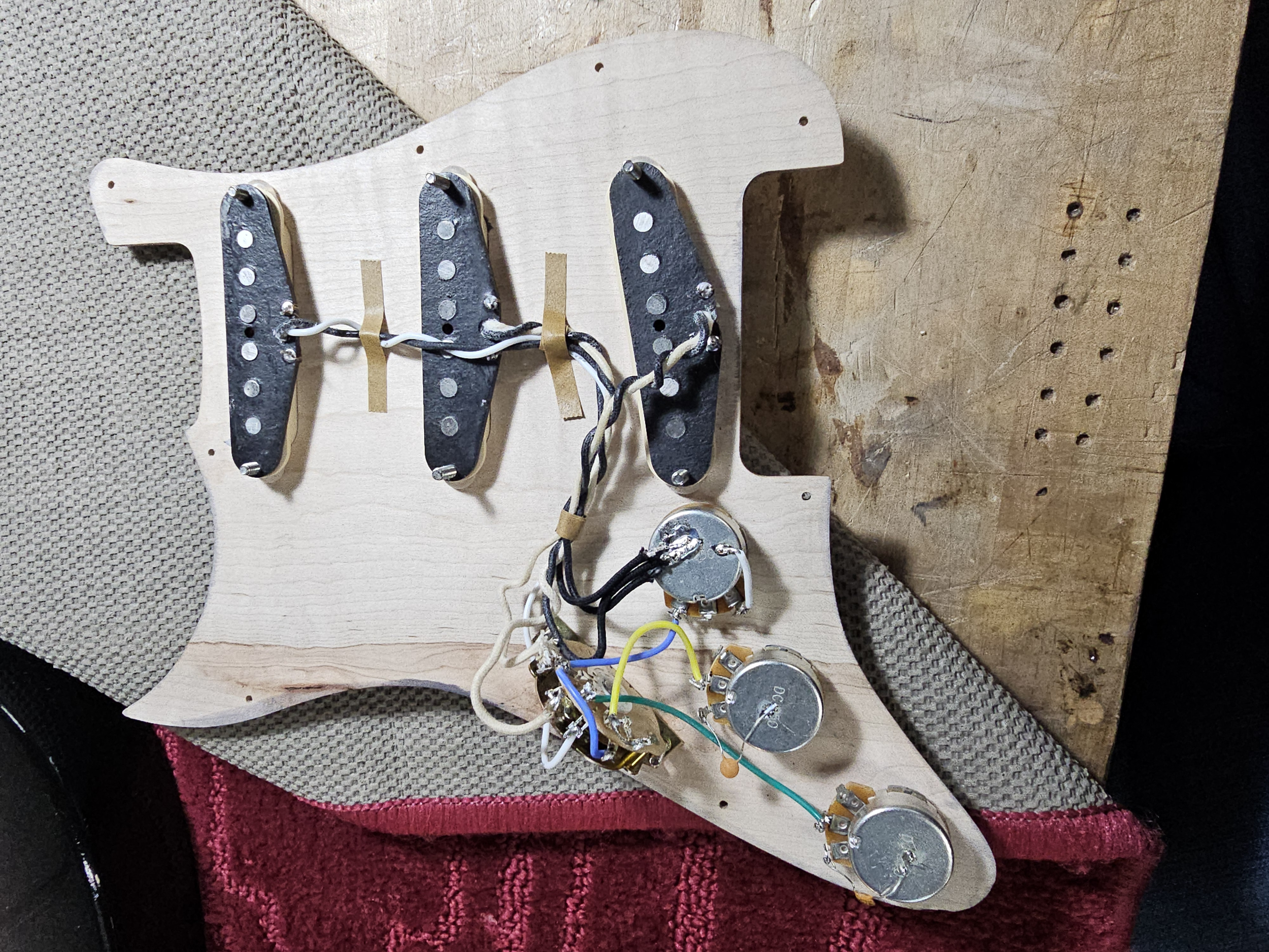An unfinished electric guitar body with pickups, wiring, and potentiometers attached, placed on a workbench with a piece of fabric underneath.