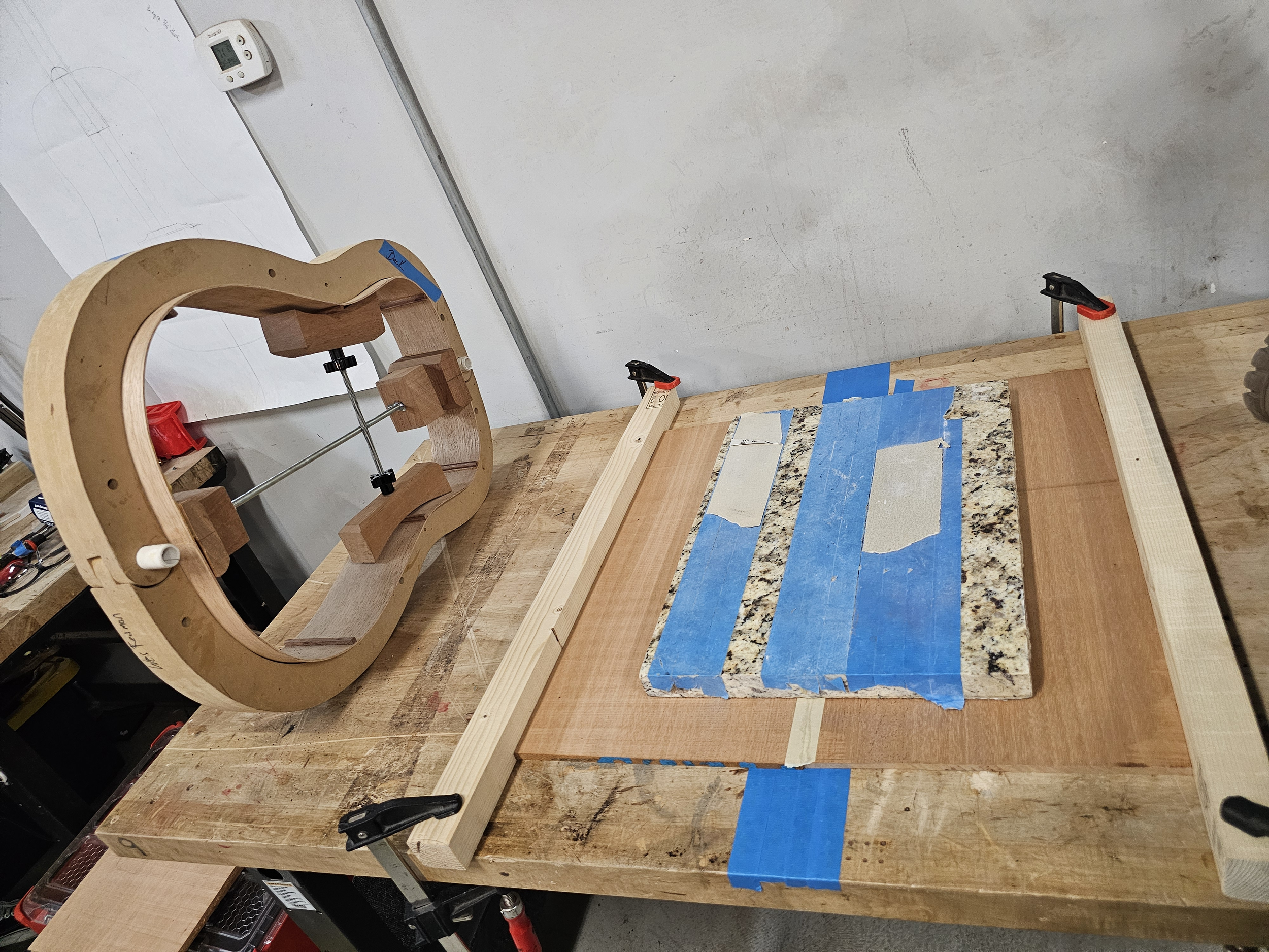 A woodworking workspace with a heart-shaped wooden frame, clamps, a piece of granite with masking tape, and a wooden board on a workbench.