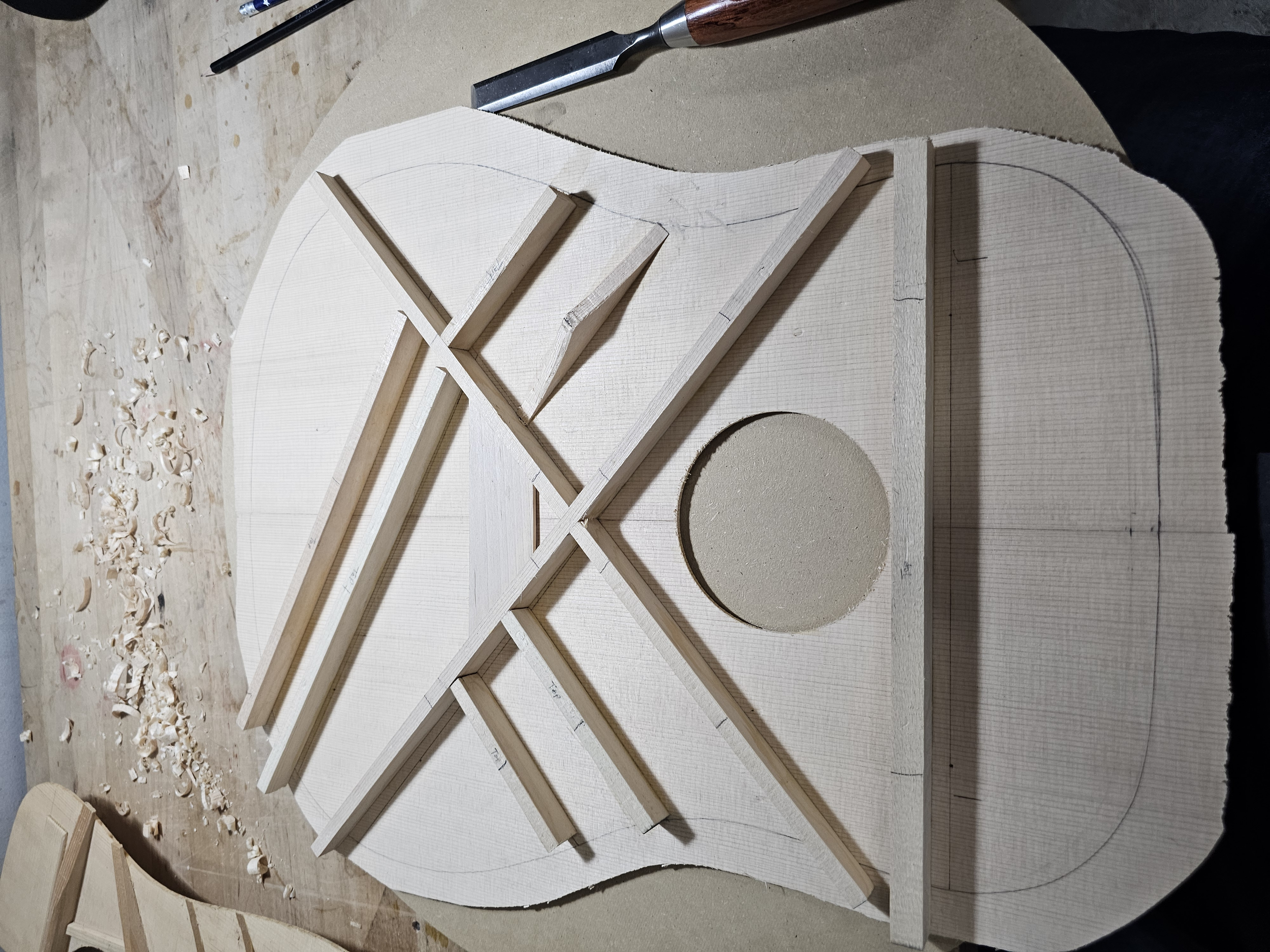 Wooden guitar body shape with fretboard and a circular sound hole cutout, resting on a workbench with wood shavings.