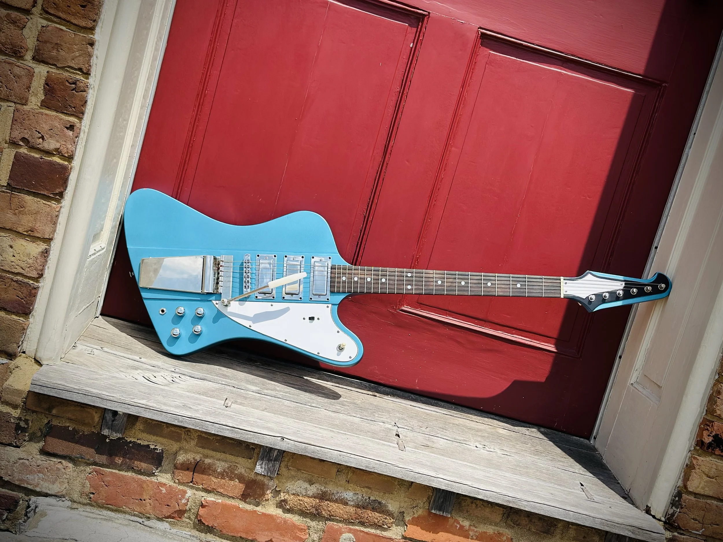 Blue electric guitar with a white pickguard leaning against a red wooden door, on a weathered wooden porch, with brick wall on the left side.