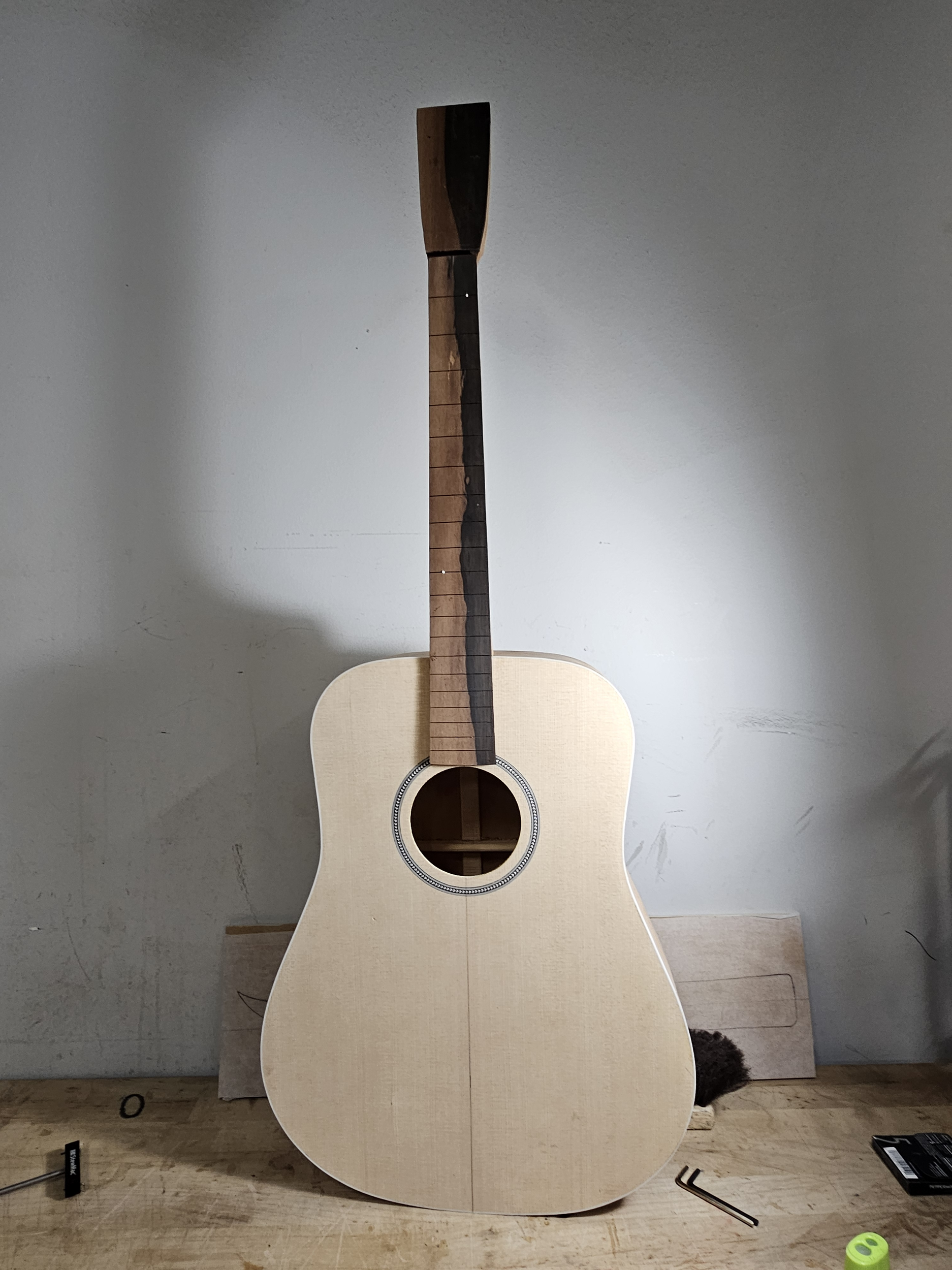 Unfinished acoustic guitar with a light-colored body and a dark neck, resting against a wall on a wooden surface.