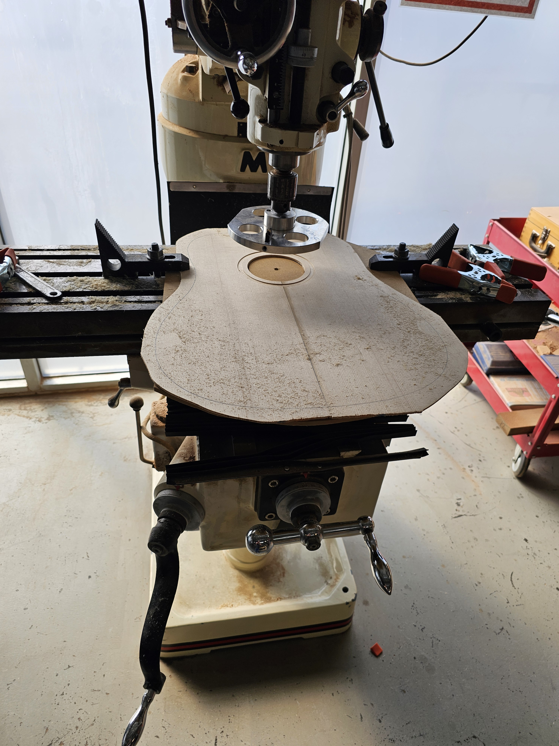 A small woodworking milling machine with a flat wooden guitar body blank mounted on it, set up for shaping or carving.