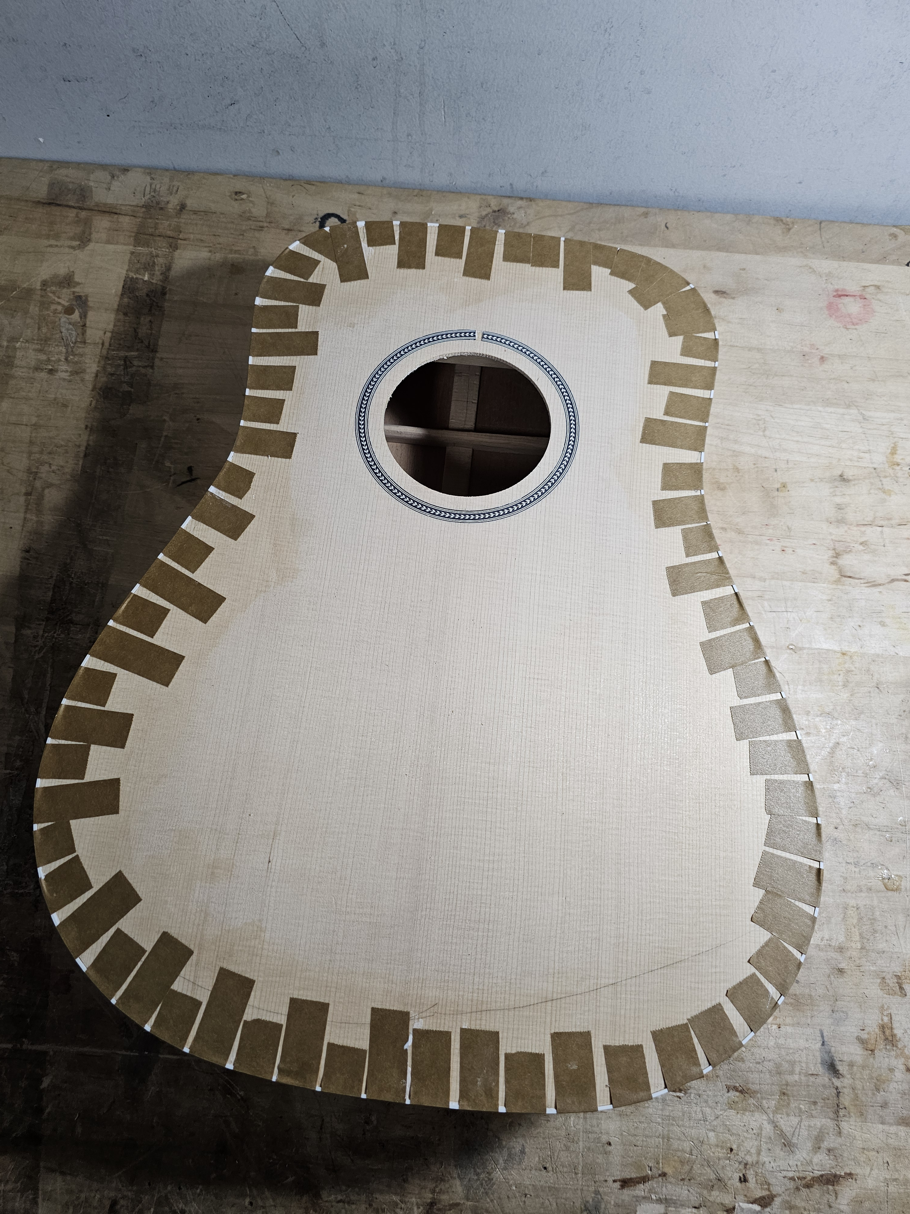 Unfinished acoustic guitar body with sound hole, laid flat on a wooden workbench.