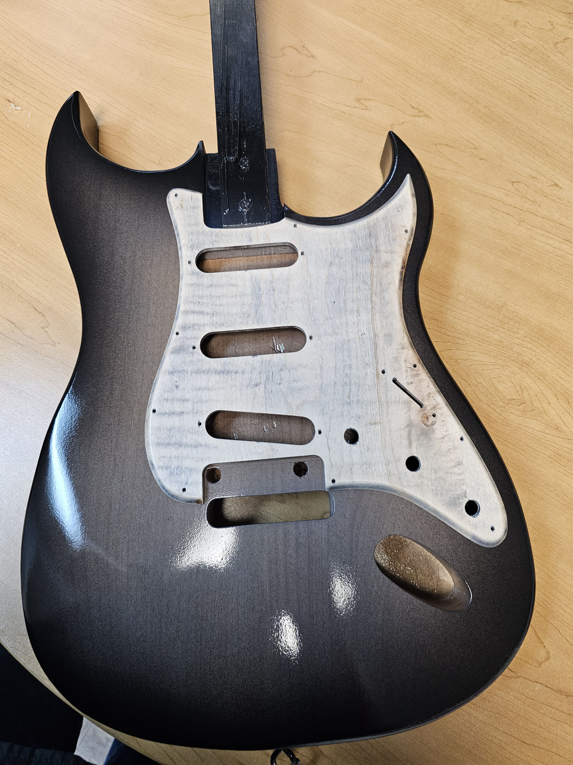 Unassembled electric guitar body with black finish and white pickguard, placed on a wooden table.