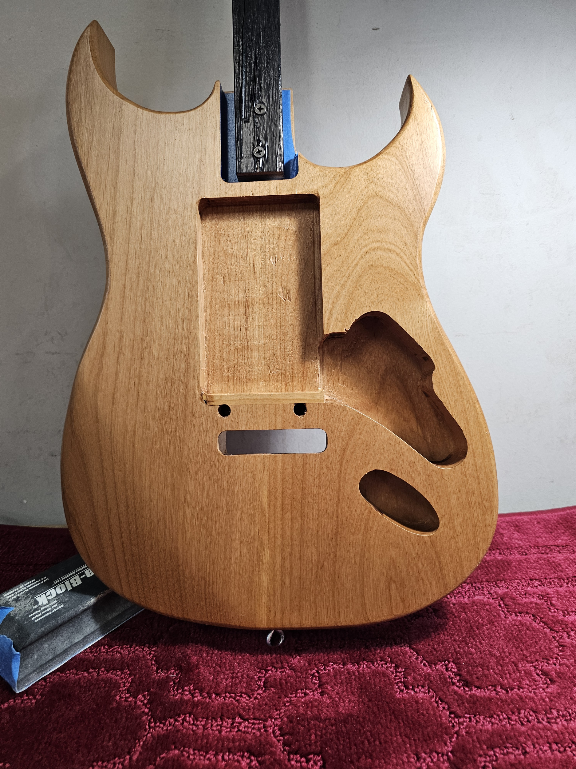 Unfinished wooden electric guitar body with cutouts for pickups and controls, on a red textured surface.