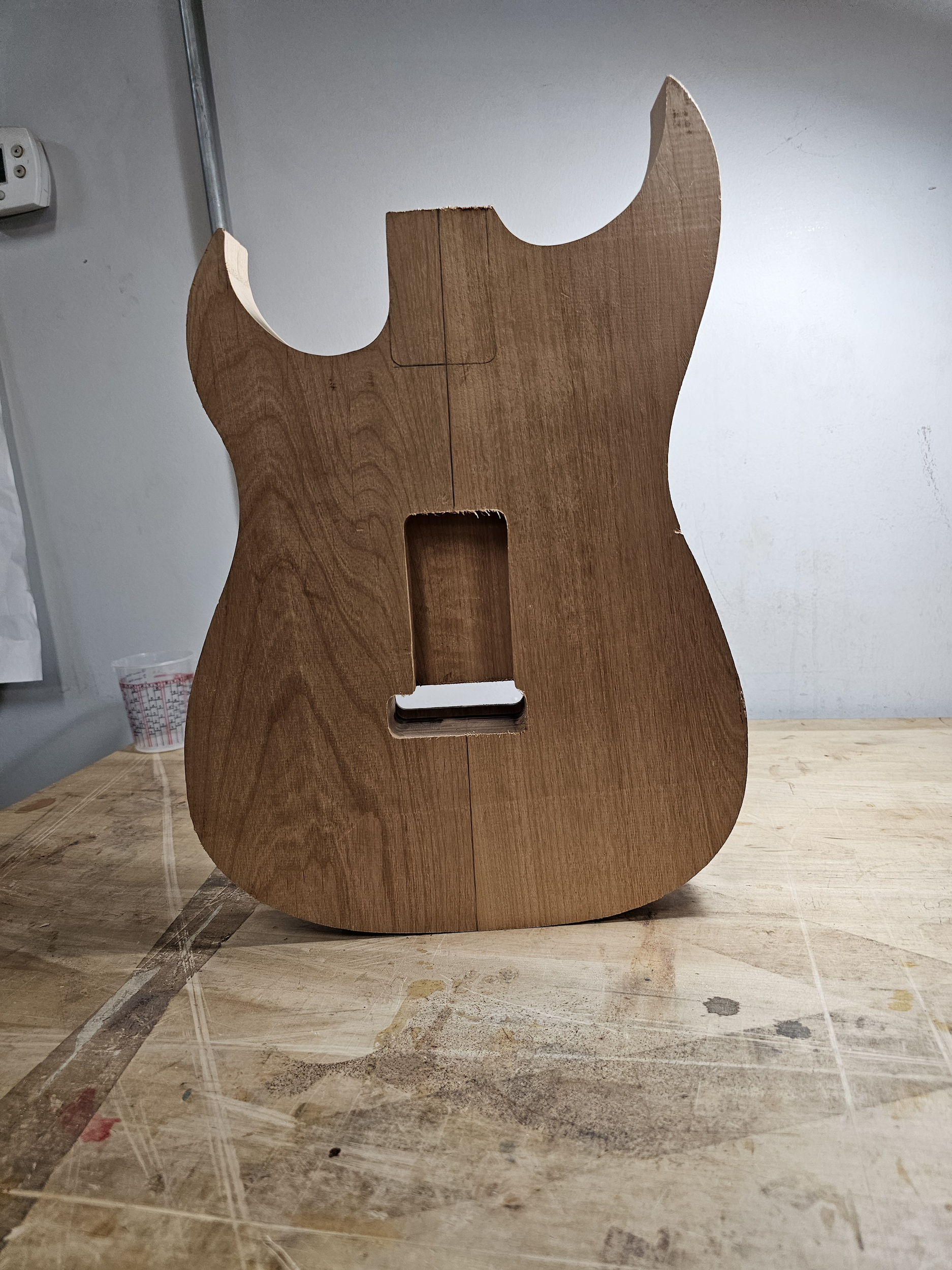 The image shows a wooden guitar body template with cutouts for the pickups and neck, placed on a worn woodworking table.